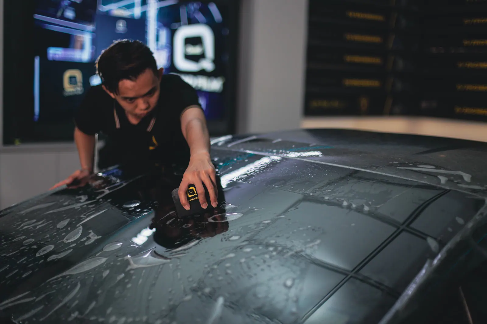 Installer working a squeegee across the roof panel of a dark car, pushing film flat over the large curved surface