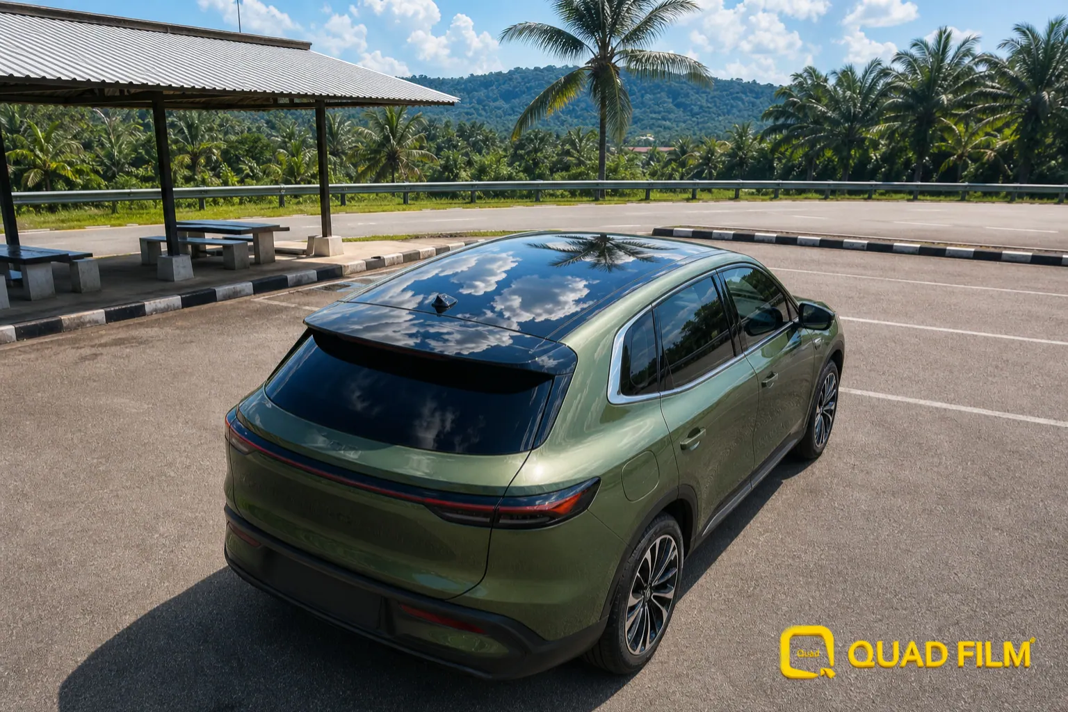 Compact green electric SUV with a panoramic glass roof parked at a Malaysian highway rest stop under midday sun